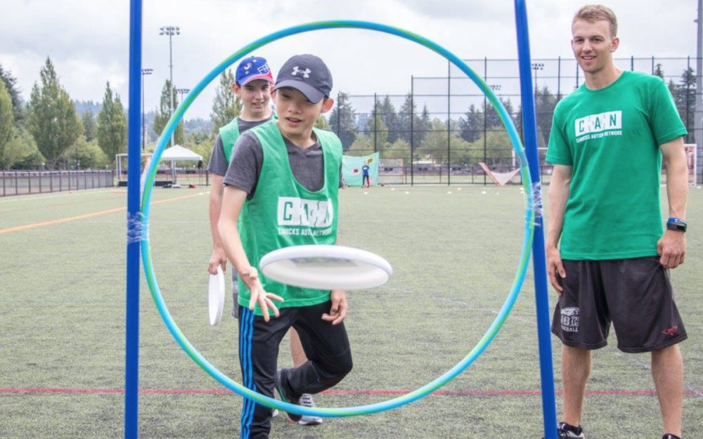 A child with autism throws a flying disc through a hoop under the guidance of a male volunteer.