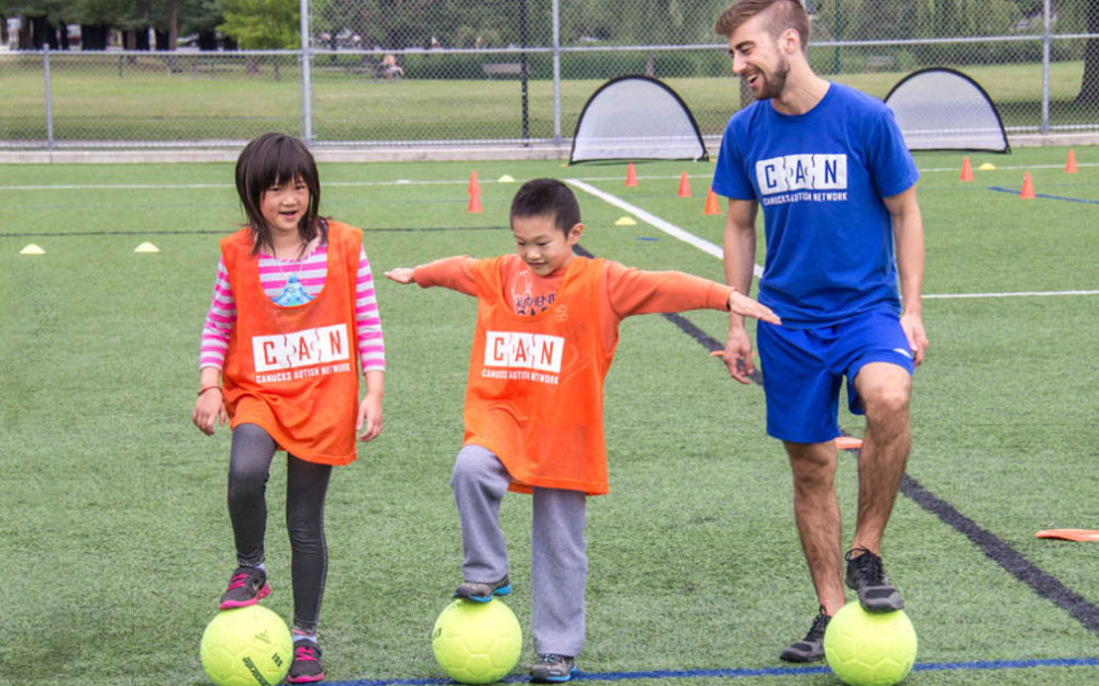 A female child, a male child and an adult male stand in a row on an outdoor turf field, each with a soccer ball under one of their feet.