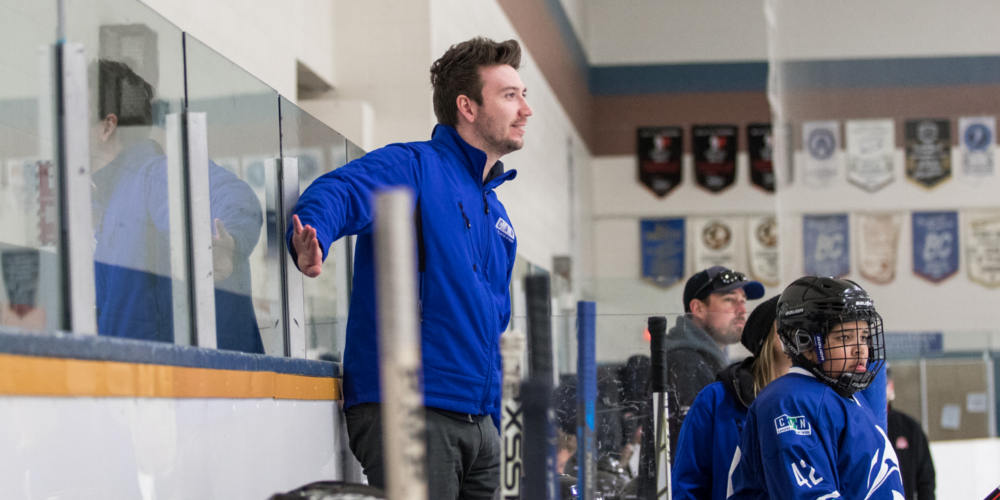 A male hockey coach stands on an arena bench behind youth hockey players during a game.